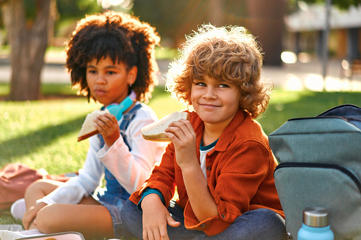 Charming schoolchildren after school resting sitting on the grass in the park. An African-American girl with a Caucasian boy took out lunchboxes with sandwiches from their backpack and eating.