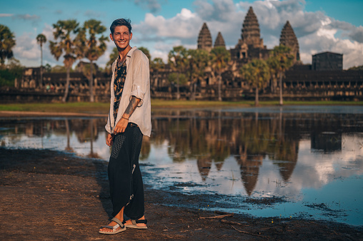 A young man standing in front of the r Angkor Wat reflecting  in the lake.