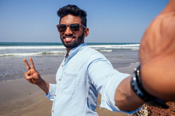 young african american man in blue shirt and jeans making a selfie portrait on the front camera of a smartphone on the ocean beach. - party beach indian ethnicity adult imagens e fotografias de stock