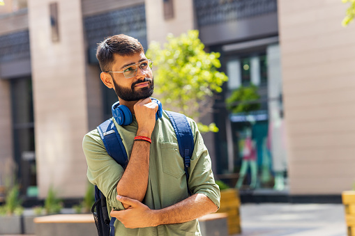 indian student with blue headset and backpack looking thoughtful at sunny day.