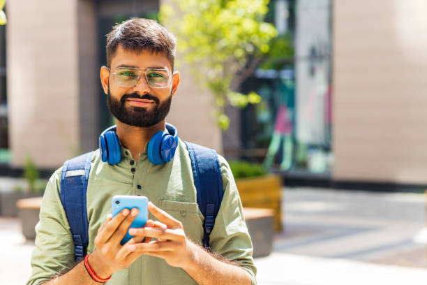 istock indian student with blue headset and backpack holding smartphone and looking at camera at sunny day 1447616565