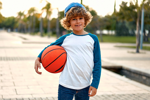 Sports and basketball. Stylish cute boy standing with a basketball ball outdoors in the park. Concept of sport, movement, healthy lifestyle, ad, action, motion.