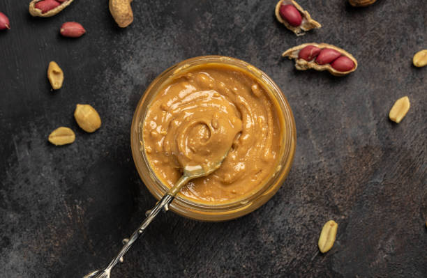 istock Peanut butter or paste in an open jar and peanuts in the peel scattered on a dark background, banner, menu, recipe place for text, top view 1447620140
