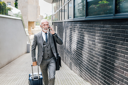 Smiling senior businessman with suitcase talking on smart phone. Elderly professional wearing suit is carrying laptop bag. He is walking on footpath by office building in the city.