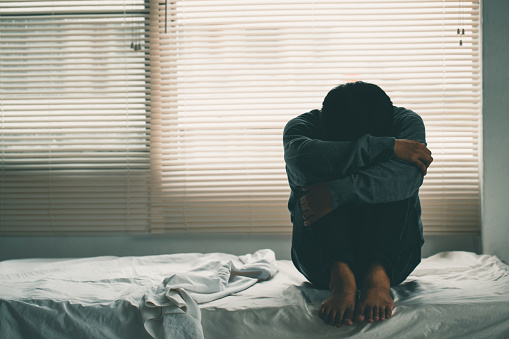 Depressed man losing his job and heartbroken sitting alone in bed near a window in dark with the low light environment, PTSD Mental health, and depression concept.psychological problem, sadness.