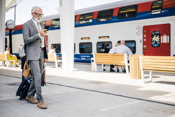 Senior male professional messaging on smart phone. Elderly businessman is walking with suitcase at subway or train station platform.