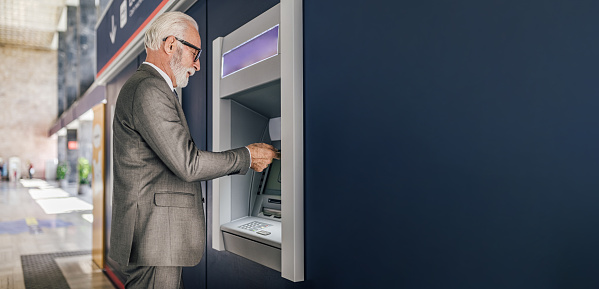 Profile view of senior businessman wearing suit using ATM machine for cash money withdrawal