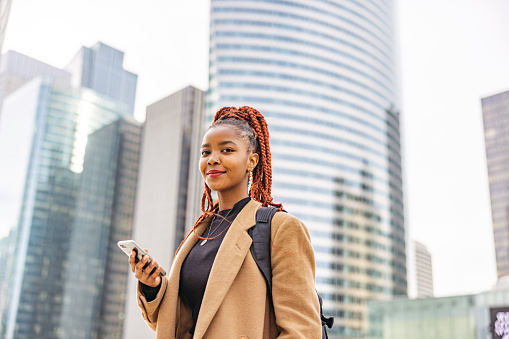 Waist up side shot from a low angle with blurred background of a young and beautiful African woman walking through the financial district of the city while holding her phone to find the address of her upcoming job interview, surrounded by skyscrapers.