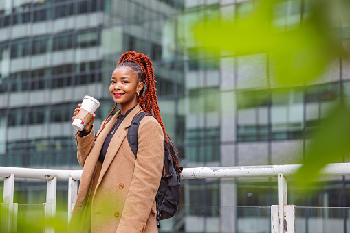 Three-quarter side shot with blurred background of a fashionable dressed beautiful African descendant woman sightseeing and holding her coffee to warm up on a cold morning in Paris.