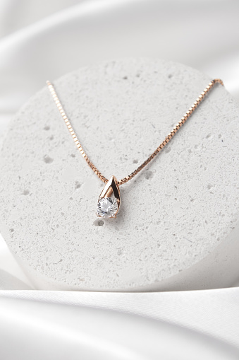 Golden necklace with crystals on concrete podium on white silk background, rose gold, single diamond pendant. Beautiful accessories for women. Elegant jewelery gift or present for wedding or saint valentine's day.