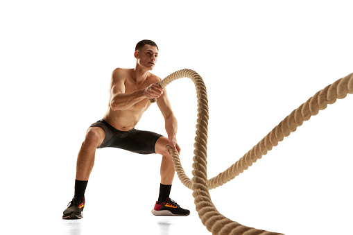 Portrait of strong young man practicing with battle ropes during workout isolated over white background. Fitness, healthy and active lifestyle concept. Shirtless athlete wearing bicycle shorts