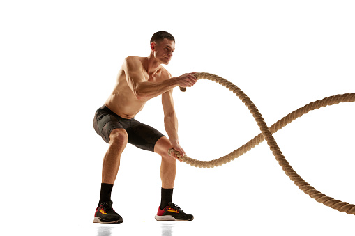 Strength. Portrait of muscular young man practicing with battle ropes during workout isolated over white background. Fitness, healthy and active lifestyle concept. Shirtless athlete in bicycle shorts