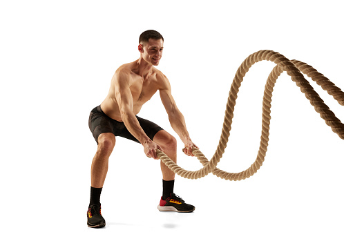 Portrait of strong young man practicing with battle ropes during workout isolated over white background. Fitness, healthy and active lifestyle concept. Shirtless athlete wearing bicycle shorts