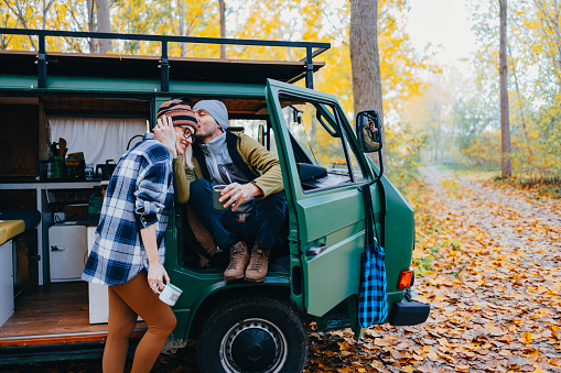 Handsome man sitting in the camping van and kissing his wife on the forehead. They are camping in the woods on an autumn day.