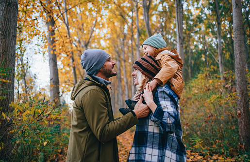 Portrait of a happy family with child taking a walk in the forest on a cold autumn day. A beautiful woman is carrying their daughter on her shoulders. They are all smiling.