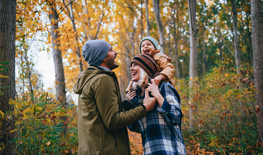 Portrait of a happy family with child taking a walk in the forest on a cold autumn day. A beautiful woman is carrying their daughter on her shoulders. They are all smiling.