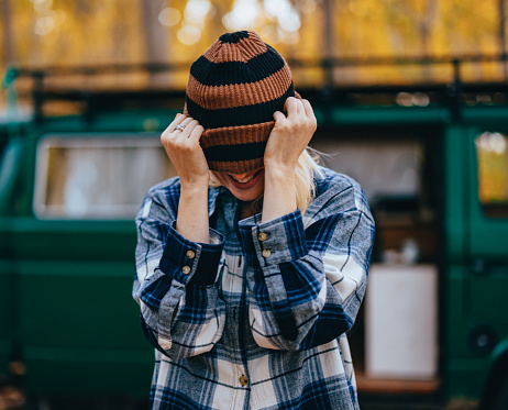 Close up shot of an anonymous woman standing in the forest in front of a camping van. She is putting a hat over her face and smiling.