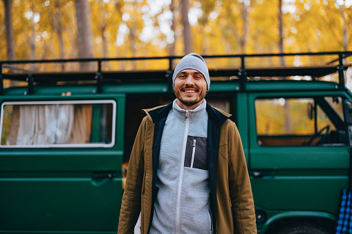 Close up shot of a handsome man wearing a hat on a cold autumn day. He is standing next to a camping van, looking at camera and smiling.