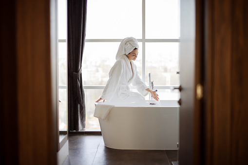 Woman wearing bathrobe sitting on edge of bathtub filling up with water