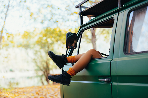 Close up shot of legs of a child hanging out and resting on the open window on a driver's seat. The van is parked in the woods in autumn.