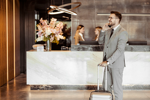 Successful good looking businessman with suitcase talking on smartphone in hotel lobby. Business trip