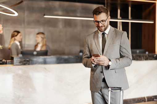 Businessman wearing formal suit holding smartphone with suitcase in hotel lobby