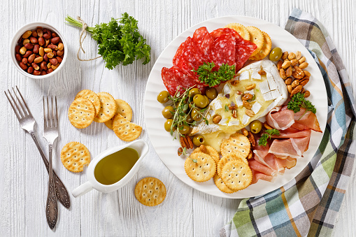 savory baked brie with crackers, spanish salchichon, salami sausages, slices of smoked ham, green olives, lettuce, peanuts, pecans on white plate on wooden table, horizontal view from above, flat lay