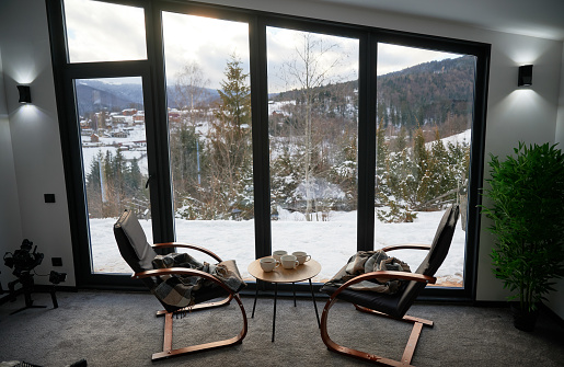 Cozy room with small coffee table and armchairs in front of large panoramic window. Front view of two wooden chairs and table against wide window with snowy mountain landscape. Concept of holidays.