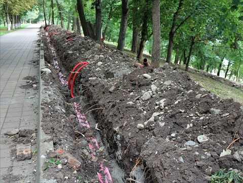 works in a city park on laying electrical communications underground, a trench along a paved path in a park with red plastic pipes with wires for installing lighting