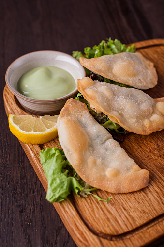 Small pasties with green salad, arugula, lemon wedges and white sauce on a wooden board. Vertical orientation