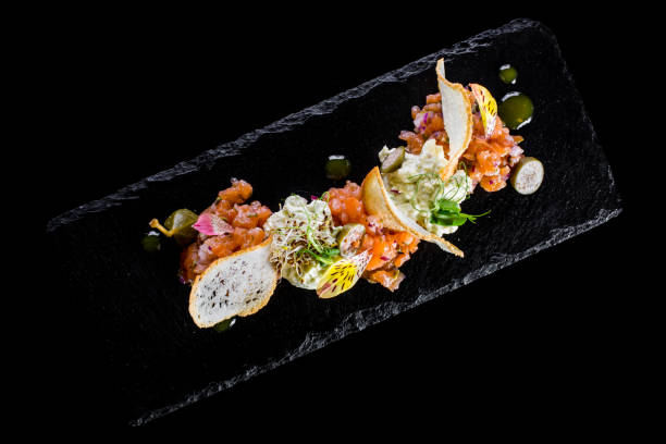 istock Salmon tartar with wheat baguette chips, microgreen lentil sprouts, capers, raspberries and edible petals. The food lies on a black slate plate, the plate lies on a black background. 1447665873