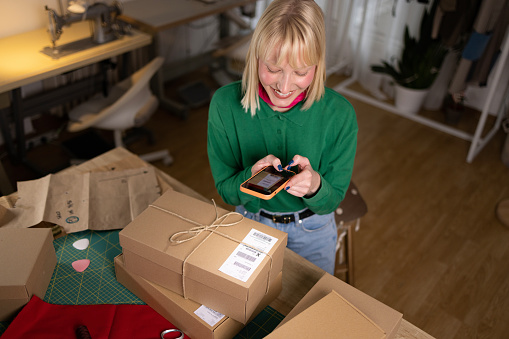 High angle view of happy female designer using smart phone while scanning bar code of a package before shipping it in a clothing design studio.