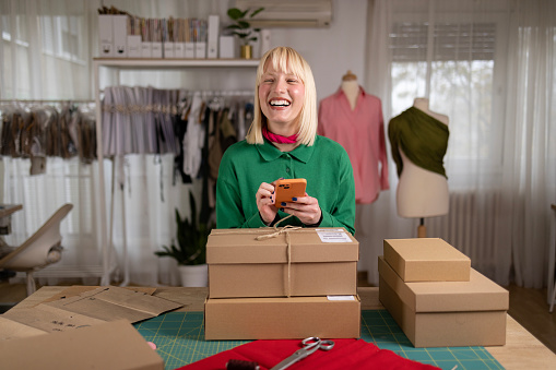 Happy fashion designer using cell phone before sending the packages in a design studio.