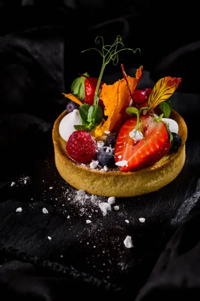 Photo of Dessert tart with strawberries, raspberries, blueberries, flower petals and powdered sugar on a stone board and a dark background. Vertical orientation