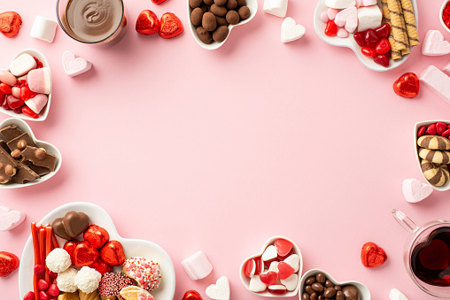 Valentine's Day concept. Top view photo of heart shaped saucers with sweets candies and glasses with drinking on isolated light pink background with empty space in the middle