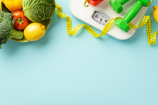 Slimming concept. Top view photo of plate with vegetables cabbage lemons cauliflower tomato dumbbells scales and tape measure on isolated pastel blue background with copyspace