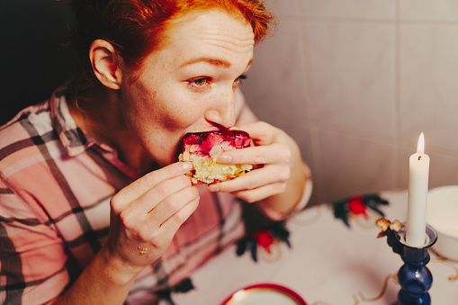 Portrait of a red-haired woman in her kitchen. She afforded herself a slice of raspberry pie. She eats the cake with her hands and bites off a big piece. It's Christmas time…