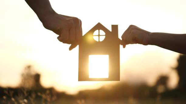 conceito de família, casa e lar felizes. mãe e filha mãos segurando uma casa de papel, casa juntos silhueta contra o pôr do sol com foco suave. conceito de família feliz, vida, seguro, cuidados - casa em exposição - fotografias e filmes do acervo