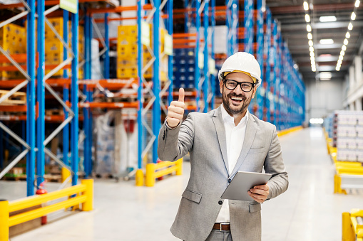 A businessman in warehouse giving thumbs up and holding tablet while smiling at the camera.