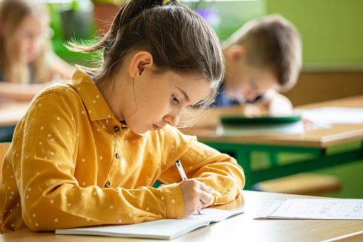 Portrait of cute girl wearing yellow shirt sitting at the desk in the classroom and writing. School children in the elementary school.