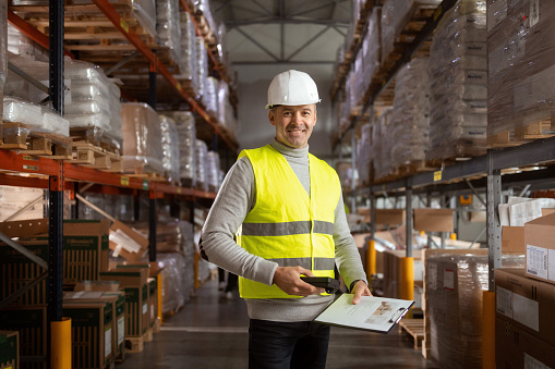 Portrait of a man working in warehouses. She is wearing protective workwear and holding a clipboard and barcode reader.
