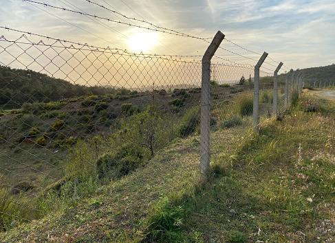 iron fence and barbed wire parallel to the dirt road. sunset in the countryside. selective focus on fence wall.