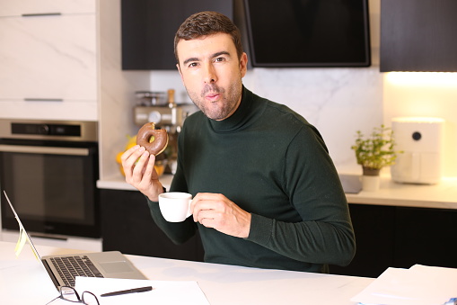 An elegant man in his forties is eating a doughnut and drinking a coffee in home office. He looks stylish is in his modern kitchen.