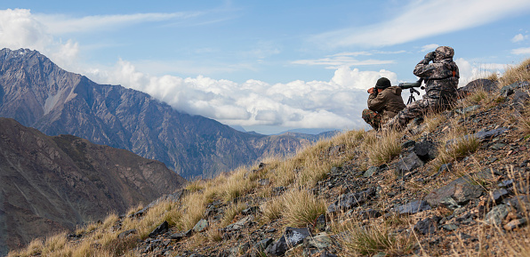 Men in camouflage with weapons are watching high in the mountains. Two hunters use optics to search for hunting objects on mountainside.
