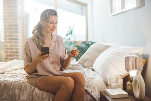 Mature woman waking up and using products for hormone replacement therapy. She is sitting on bed in pajamas and reading instructions on smart phone.