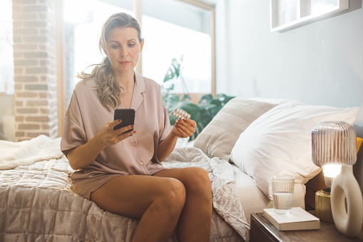 Mature woman waking up and using products for hormone replacement therapy. She is sitting on bed in pajamas and reading instructions on smart phone.
