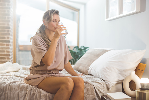Mature woman waking up and using products for hormone replacement therapy. She is sitting on bed in pajamas and drinking water.