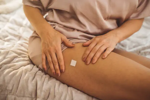 Mature woman waking up and using products for hormone replacement therapy. She is sitting on bed in pajamas and putting adhesive patch on leg.