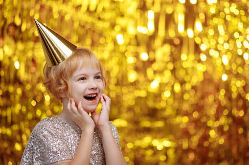 A child in a festive hat and a shiny dress is fooling around at a party on a golden background. Holiday concept, congratulations. Place for your text. High quality photo