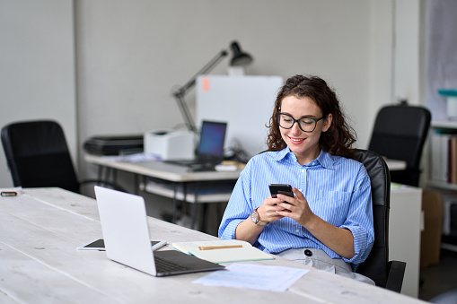 Young happy business woman entrepreneur looking at smartphone using cellphone mobile cell technology, professional businesswoman manager working in office typing on mobile cell phone sitting at desk.